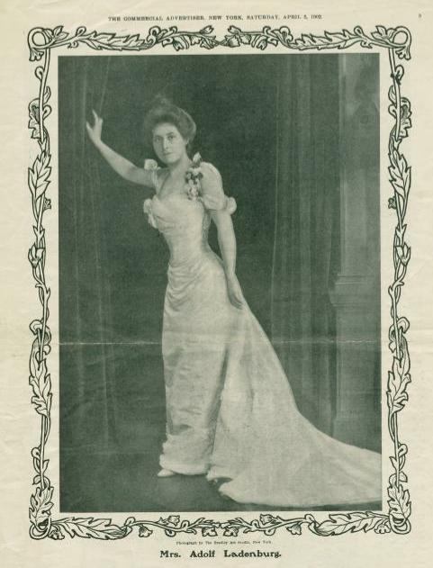 Black and white full-length photo of Emily Ladenburg. She is wearing a white  full-length gown with a train at the back. Flowers are arranged at the shoulders of the gown. Her hair is dressed in a knot at the top of head in the style popular for the time. 